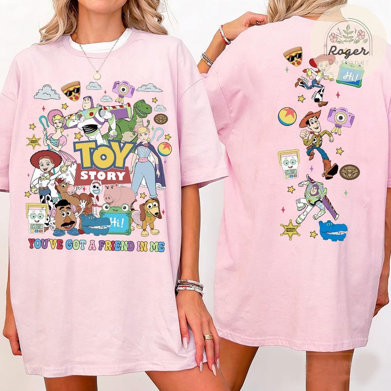 May include: A light pink t-shirt featuring a colorful Toy Story graphic on the front and back. The front displays various characters, the Toy Story logo, and the text You've Got A Friend In Me. The back shows smaller character icons and motifs.