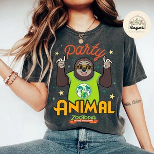 May include: Dark gray t-shirt featuring a cartoon sloth wearing sunglasses and a green shirt with the text "Party Animal" in orange and yellow. The shirt also has the text "Zootopia Better Together!" and star graphics.