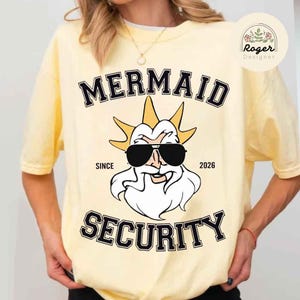 May include: A pale yellow t-shirt with the text "MERMAID SECURITY" in black, block lettering. The design includes a cartoon illustration of a bearded figure wearing sunglasses and a crown. The words "SINCE 2026" are also present.
