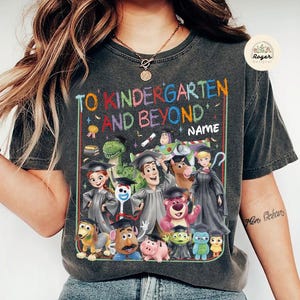 Peut inclure: T-shirt gris fonc&eacute; avec l'inscription "TO KINDERGARTEN AND BEYOND NAME" en lettres color&eacute;es. Comporte un graphisme de divers personnages de dessins anim&eacute;s portant des chapeaux de remise de dipl&ocirc;mes. Le t-shirt a un aspect vintage et us&eacute;.