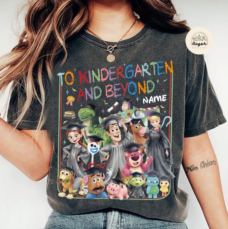 Peut inclure: T-shirt gris fonc&eacute; avec l'inscription "TO KINDERGARTEN AND BEYOND NAME" en lettres color&eacute;es. Comporte un graphisme de divers personnages de dessins anim&eacute;s portant des chapeaux de remise de dipl&ocirc;mes. Le t-shirt a un aspect vintage et us&eacute;.