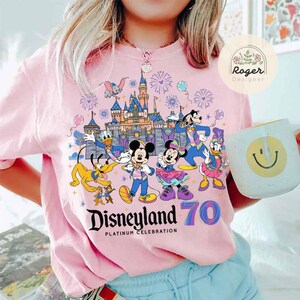 K&ouml;nnte beinhalten: Pinkes T-Shirt mit einer bunten Grafik von Mickey Mouse und seinen Freunden, die das 70-j&auml;hrige Jubil&auml;um von Disneyland feiern. Die Grafik zeigt Mickey Mouse, Minnie Mouse, Goofy, Donald Duck, Pluto und das Disneyland-Schloss. Der Text "Disneyland 70 Platinum Celebration" ist unter der Grafik aufgedruckt.