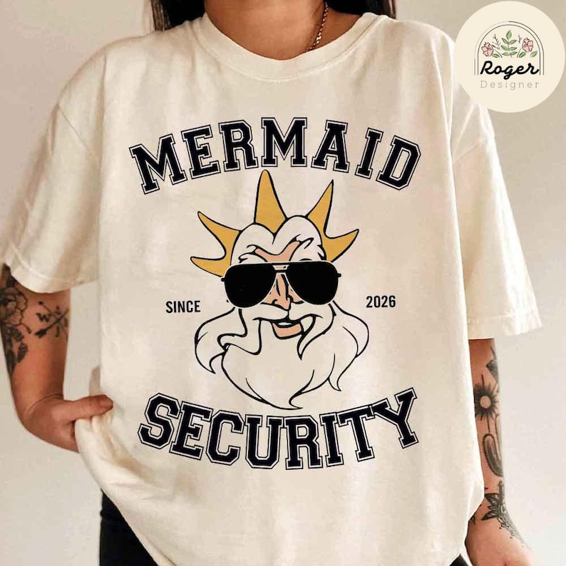 May include: A cream-coloured t-shirt featuring the text "MERMAID SECURITY" in black. Below the text is a cartoon illustration of a bearded figure wearing sunglasses. The shirt also includes the text "SINCE 2026".
