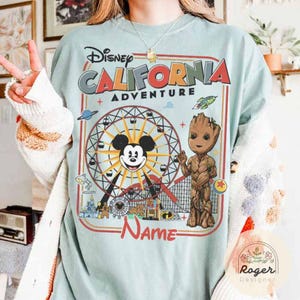 May include: Light blue t-shirt featuring a colorful graphic of the Disney California Adventure theme park. The design includes Mickey Mouse, Groot, a Ferris wheel, and the text "California Adventure". The shirt has a vintage, distressed look.