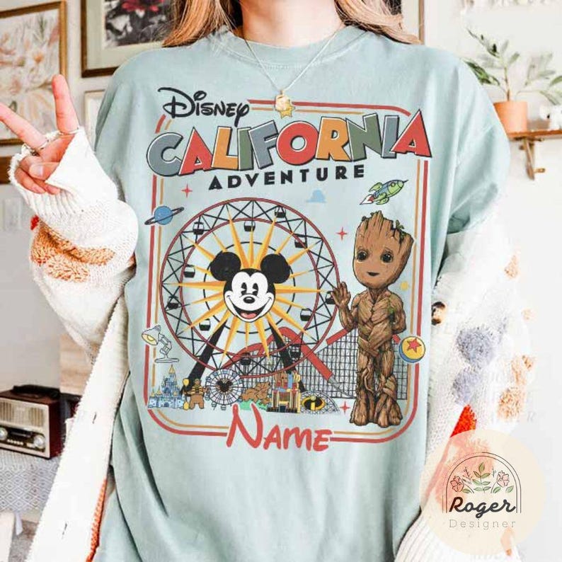 May include: Light blue t-shirt featuring a colorful graphic of the Disney California Adventure theme park. The design includes Mickey Mouse, Groot, a Ferris wheel, and the text "California Adventure". The shirt has a vintage, distressed look.