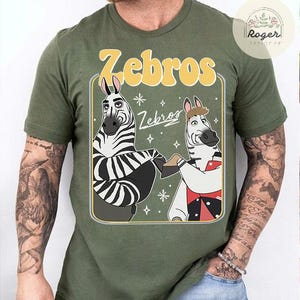 Pu&ograve; includere: T-shirt verde oliva con un design a cartoni animati di due zebre che si tengono per mano. Il design include la parola "Zebros" in un font retr&ograve; giallo e la parola "Zebros" in un font pi&ugrave; piccolo. Le zebre sono bianche e nere.