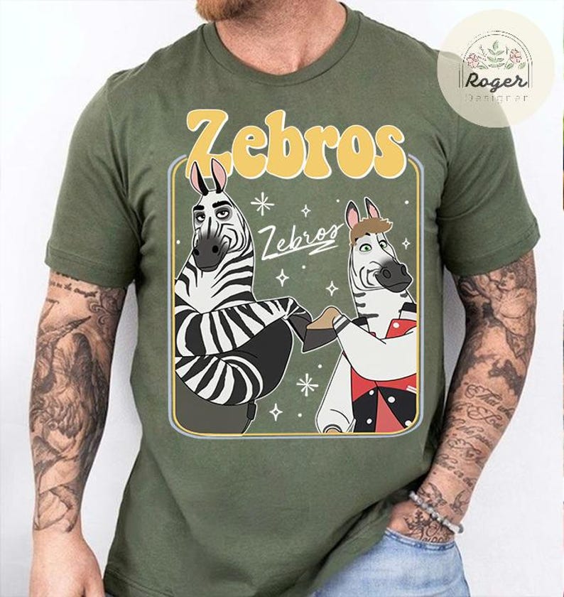 Pu&ograve; includere: T-shirt verde oliva con un design a cartoni animati di due zebre che si tengono per mano. Il design include la parola "Zebros" in un font retr&ograve; giallo e la parola "Zebros" in un font pi&ugrave; piccolo. Le zebre sono bianche e nere.