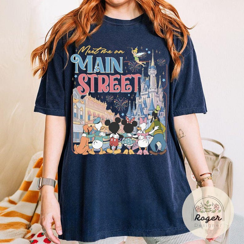 May include: Navy blue t-shirt featuring the text "Meet me on Main Street" above a graphic of Disney characters and Cinderella's castle. The graphic includes Mickey and Minnie Mouse, Goofy, Donald and Daisy Duck.