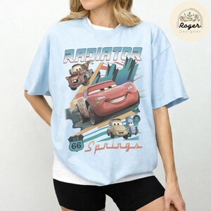May include: A light blue t-shirt featuring a graphic print of characters from the movie Cars, including Lightning McQueen, Mater, Luigi, and Guido. The design includes the text Radiator Springs and a US 66 road sign emblem on the front.