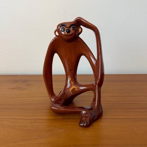 May include: A brown ceramic monkey figurine in a seated pose, with one arm raised to its head. The monkey has a smooth, glossy finish and black details for the eyes and ears. The figurine is displayed on a wooden surface.