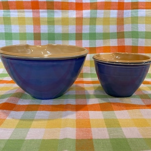 以下が含まれることがあります： 茶色の縁取りのある青いセラミック製のボウルが2つ。大きなボウルは奥に、小さなボウルは手前にあります。両方のボウルはチェック柄のテーブルクロスの上に置かれています。