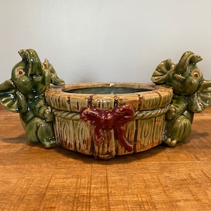 May include: A ceramic planter featuring two green elephants supporting a wooden-style basket. The basket has a red bow and rope detailing. The elephants have their trunks raised. The planter is suitable for small plants or decorative items.