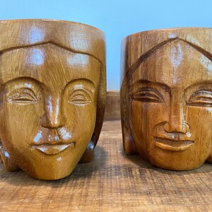 May include: Two wooden Buddha face planters. The planters are carved from light brown wood and feature detailed facial features, including closed eyes and serene expressions. The planters are displayed on a wooden surface.