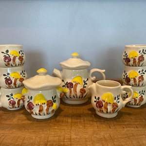 May include: A vintage mushroom-themed tea set with a teapot, creamer, sugar bowl, and four cups. The set is decorated with yellow and brown mushrooms on a cream-colored background.