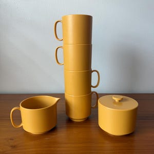 May include: A set of mustard-yellow kitchenware, including four stacked mugs, a creamer, and a sugar bowl with a lid. The mugs have curved handles. The items are arranged on a wooden surface against a white wall.
