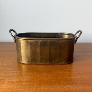 May include: A vintage brass planter with a rectangular shape and two curved handles. The planter has vertical striped detailing and a warm, aged patina. It sits on a wooden surface, suggesting a decorative or functional item for home decor.