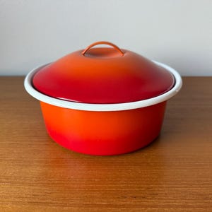 Może przedstawiać: Okrągły, pomarańczowo-czerwony garnek emaliowany z pokrywką. Garnek ma białą obwódkę i mały uchwyt na pokrywce. Garnek stoi na drewnianej powierzchni. Naczynia kuchenne.
