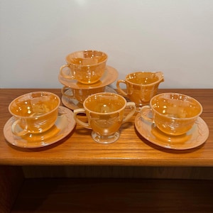May include: A set of iridescent orange teacups, saucers, and a creamer on a wooden surface. The teacups and saucers have a floral design. The creamer has a handle and a spout. The set is arranged on a wooden table.