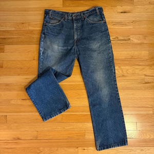 May include: Blue denim jeans with a classic five-pocket design. The jeans have a straight leg cut and a button and zip closure. The denim has a faded wash, giving them a vintage look. The jeans are laid flat on a wooden surface.
