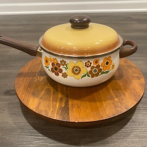 May include: A vintage yellow and brown enamel saucepan with a floral design. The saucepan has a brown handle and a brown lid with a knob. The saucepan is sitting on a wooden trivet.