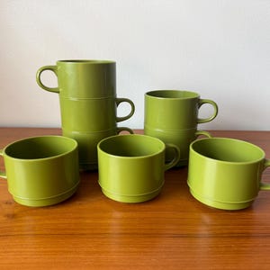 以下が含まれることがあります： 丸みを帯びた底と湾曲したハンドルが付いた、6つの緑色のマグカップのセット。 マグカップは2つと3つのグループに積み重ねられ、3つのマグカップが個別に配置されています。 マグカップは、無地の不透明な素材で作られています。