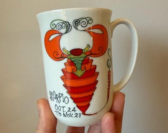 May include: White ceramic mug featuring a colorful illustration of the Scorpio zodiac sign. The design includes orange, red, and green elements, with the text "SCORPIO OCT. 24 TO NOV. 21". The mug has a curved handle.
