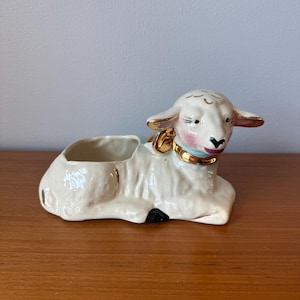 May include: A ceramic lamb planter in cream with gold accents. The lamb is lying down with a small opening for planting. The lamb has pink cheeks, a black nose, and a gold collar. The planter is a decorative item.