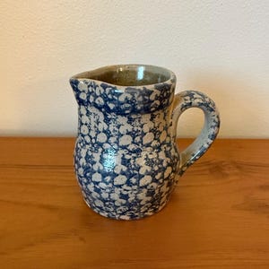 May include: A small ceramic jug with a speckled blue and white pattern. The jug has a handle and a spout. The interior is light brown. The jug is on a wooden surface.
