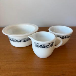 Puede incluir: Un juego de fuentes de cerámica blanca con un diseño floral azul. El conjunto incluye una jarrita, un cuenco y una taza pequeña. Los platos están colocados sobre una superficie de madera.