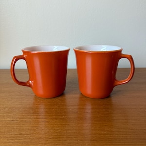 Puede incluir: Dos tazas de cerámica naranja con interiores blancos se exhiben sobre una superficie de madera. Las tazas tienen un diseño simple y redondeado con un asa en el lateral. Las tazas están vacías y listas para ser llenadas con una bebida.