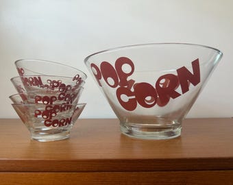 Op de afbeelding: Een set heldere glazen popcornkommen. Eén grote kom en vier kleinere kommen zijn zichtbaar. Het woord "POP CORN" is in het rood op de kommen gedrukt. De kommen staan op een houten oppervlak.
