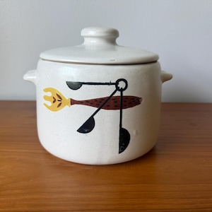 Peut inclure: Un pot en céramique crème avec couvercle, finition mouchetée et deux anses latérales. L'avant présente une illustration graphique d'une fourchette jaune et de trois cuillères à mesurer noires reliées par des lignes fines sur un manche brun.