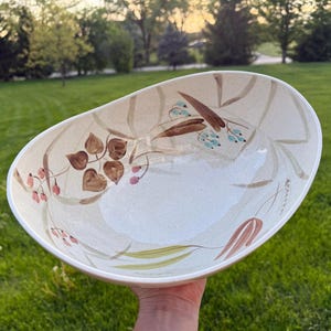 May include: Oval-shaped ceramic serving bowl with a cream-colored base. The bowl is decorated with hand-painted botanical designs in shades of brown, pink, and teal. The bowl is held in front of a green grassy background.