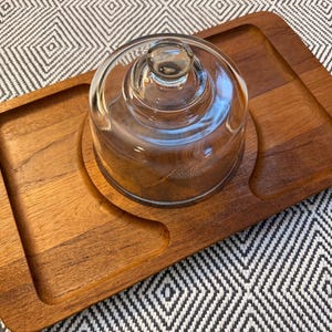 Puede incluir: Una bandeja de madera para servir con una cúpula de cristal transparente. La bandeja es rectangular con esquinas redondeadas y un área empotrada para la cúpula. La madera tiene un acabado cálido y natural. La cúpula de cristal tiene un asa pequeña en la parte superior.