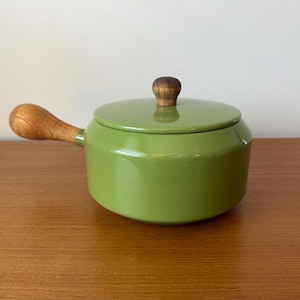 May include: A small, green enameled saucepan with a wooden handle and lid. The lid has a wooden knob. The saucepan is sitting on a wooden surface. The handle is curved and attached to the side of the pan.