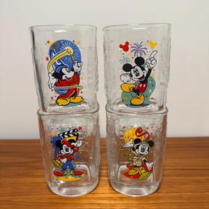 Puede incluir: Cuatro vasos de vidrio transparente con ilustraciones de Mickey Mouse. Cada vaso tiene un diseño diferente de Mickey, con fuegos artificiales y el año 2000 impreso en algunos. Los vasos están apilados sobre una superficie de madera.