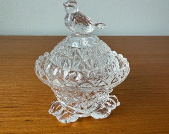 May include: A clear glass trinket box with a bird-shaped lid. The box has a round, faceted design and sits on a base with decorative feet. The bird on top is also clear and detailed.