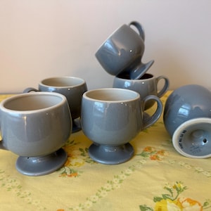 May include: A collection of six gray ceramic mugs with rounded bodies and small handles. The mugs have a pedestal base and a light-colored interior. One mug is upside down, revealing the base.