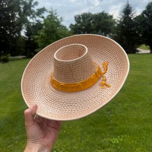 Könnte beinhalten: Eine Servierschale aus Keramik in Form eines Sombreros mit hellbeigem, texturiertem Flechtmuster und einem leuchtend gelben Zierband mit Quastendetail. Die Schale hat eine breite Krempe und eine erhöhte mittlere Krone zum Servieren.