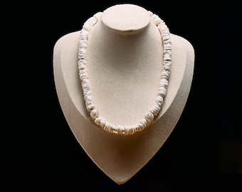 Op de afbeelding: Een witte schelpenketting tentoongesteld op een crèmekleurige buste tegen een zwarte achtergrond. De ketting is samengesteld uit kleine, onregelmatig gevormde schelpstukken die aan elkaar zijn geregen. De algehele esthetiek is eenvoudig en natuurlijk.