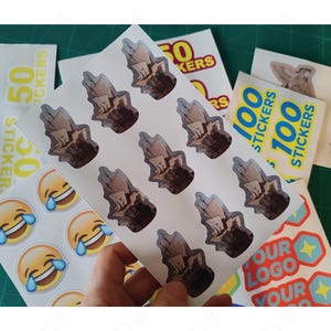 May include: Sheets of stickers featuring various designs. One sheet displays multiple stickers with a brown and beige illustration. Other sheets show stickers with the text "50 STICKERS" and "100 STICKERS". Additional stickers have a laughing emoji and a logo design.