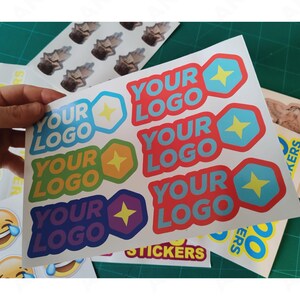 May include: A sheet of colorful stickers with the text "YOUR LOGO" in various colors, each with a star and geometric shape. Other stickers include a laughing emoji and small images. The stickers are on a green cutting mat.