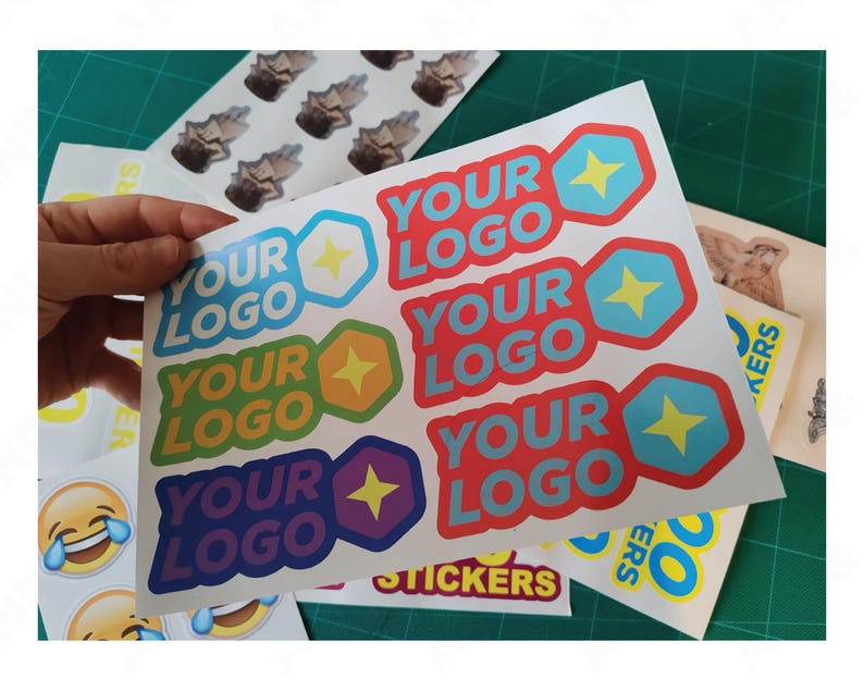May include: A sheet of colorful stickers with the text "YOUR LOGO" in various colors, each with a star and geometric shape. Other stickers include a laughing emoji and small images. The stickers are on a green cutting mat.