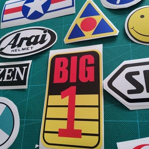May include: A selection of stickers with diverse designs and text, arranged on a green surface. Stickers include the text "BIG 1", "Arai HELMET", and a smiley face. Other designs include a star, a triangle, and a circle.