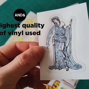 May include: A sticker featuring a classical figure in a flowing blue gown with intricate details. The sticker is held by a hand, with the text "AND&" and "Highest quality of vinyl used" visible.
