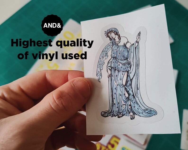 May include: A sticker featuring a classical figure in a flowing blue gown with intricate details. The sticker is held by a hand, with the text "AND&" and "Highest quality of vinyl used" visible.