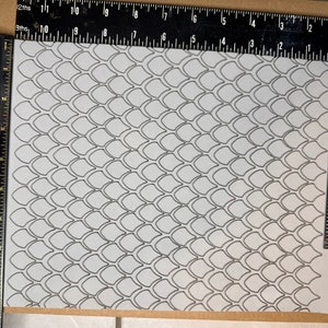 Op de afbeelding: Een wit vel papier met een zwart-wit patroon van overlappende schubben. Het papier ligt op een bruin oppervlak met een zwarte liniaal aan de linker- en rechterkant.