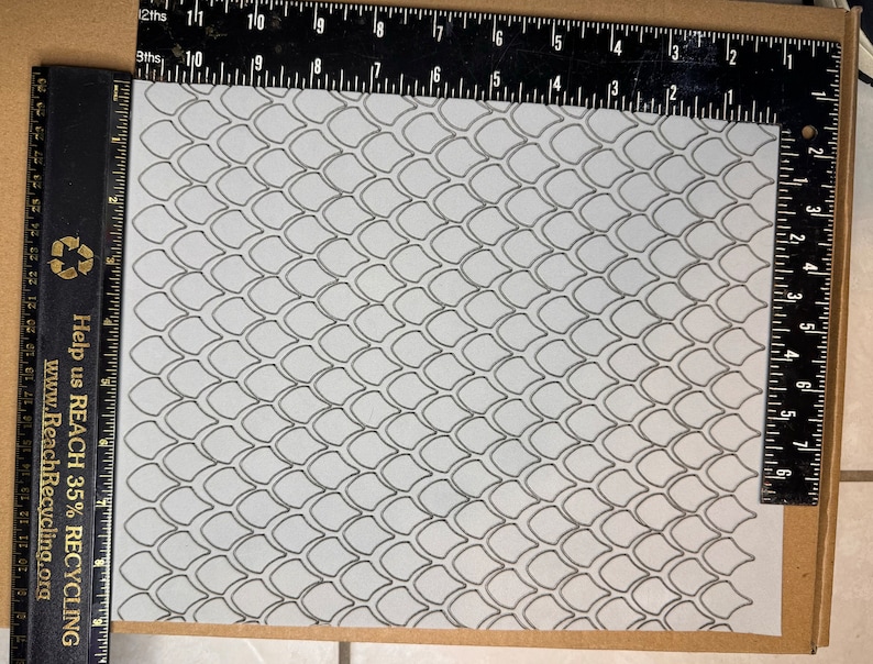 Op de afbeelding: Een wit vel papier met een zwart-wit patroon van overlappende schubben. Het papier ligt op een bruin oppervlak met een zwarte liniaal aan de linker- en rechterkant.