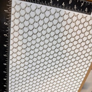 Op de afbeelding: Een close-up van een wit, getextureerd vel met een herhalend patroon van kleine, verhoogde, zilverkleurige schubben. Het vel wordt gemeten met een liniaal in inches.