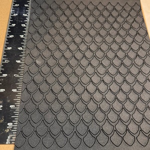 Op de afbeelding: Zwarte, getextureerde plaat met een herhalend patroon van verhoogde, overlappende schubben. De schubben zijn in een verspringend patroon gerangschikt, waardoor een subtiel 3D-effect ontstaat.